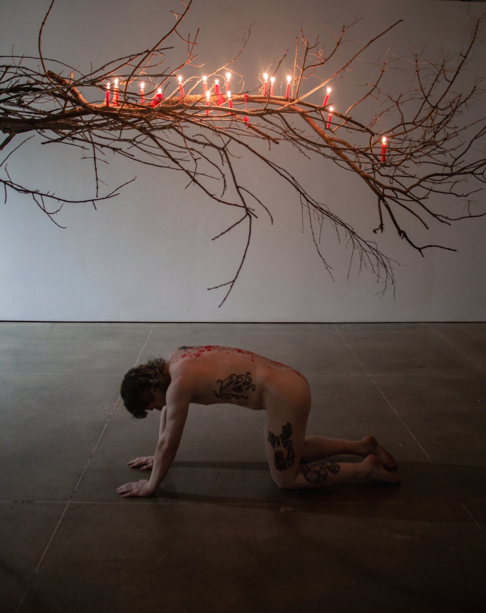 Swan Ferraro installation image 6 - a person on the ground in a dark room, underneath a tree branch covered in lit candles. Wax is dripping onto the person